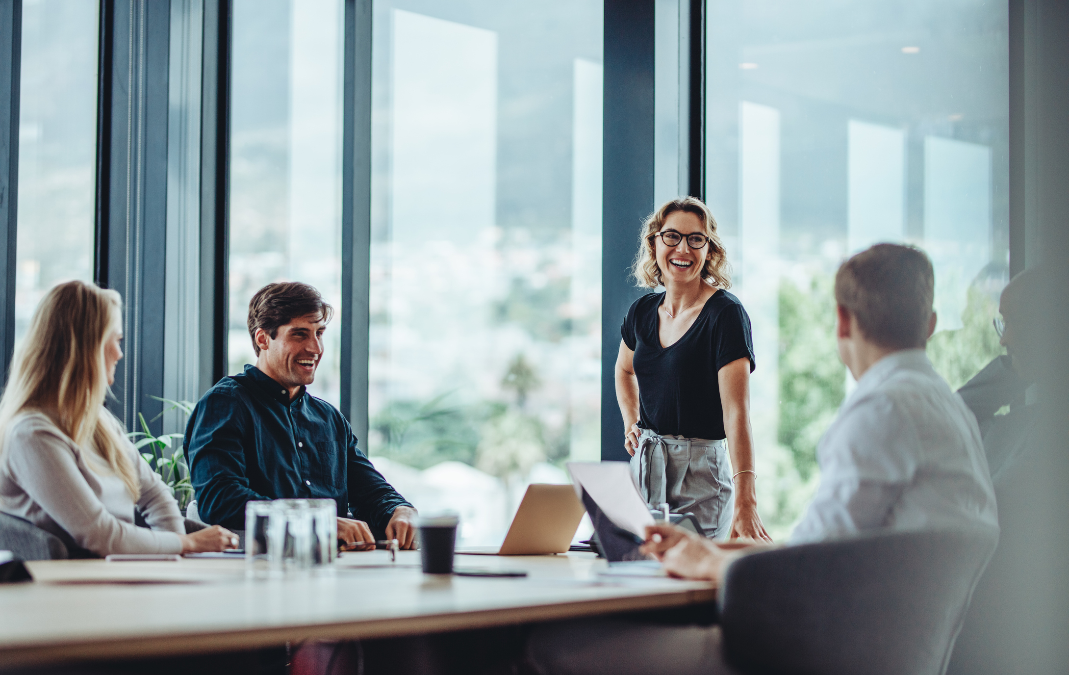 A team of professionals in a meeting, with a woman standing and smiling at her colleagues.